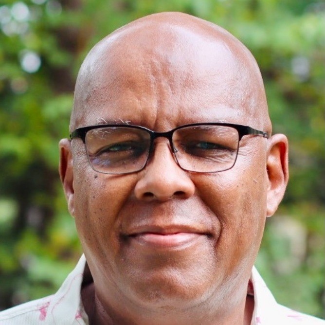 A headshot of a man wearing glasses with a slight smile and a blurred background of greenery.